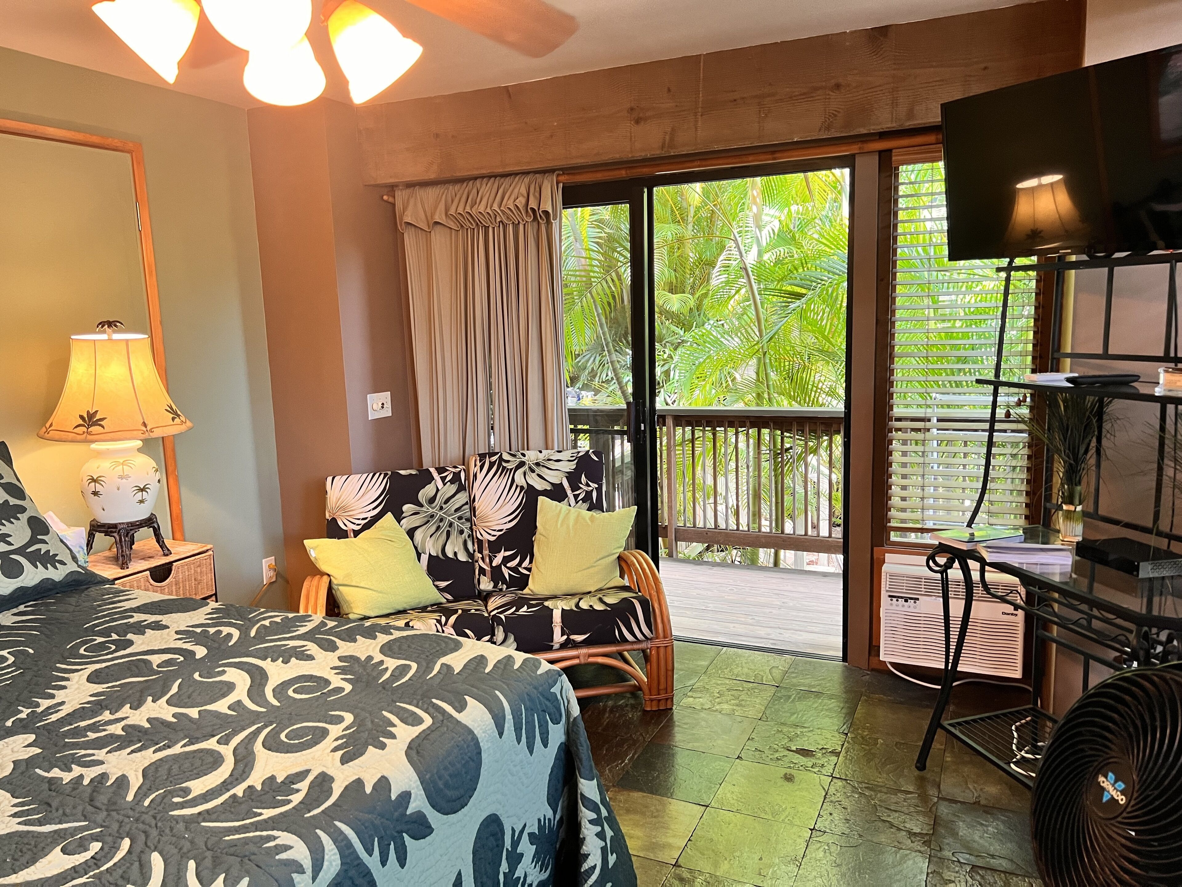 A hotel room with a bed, a sofa, a TV, a lamp, and a view of greenery outside.