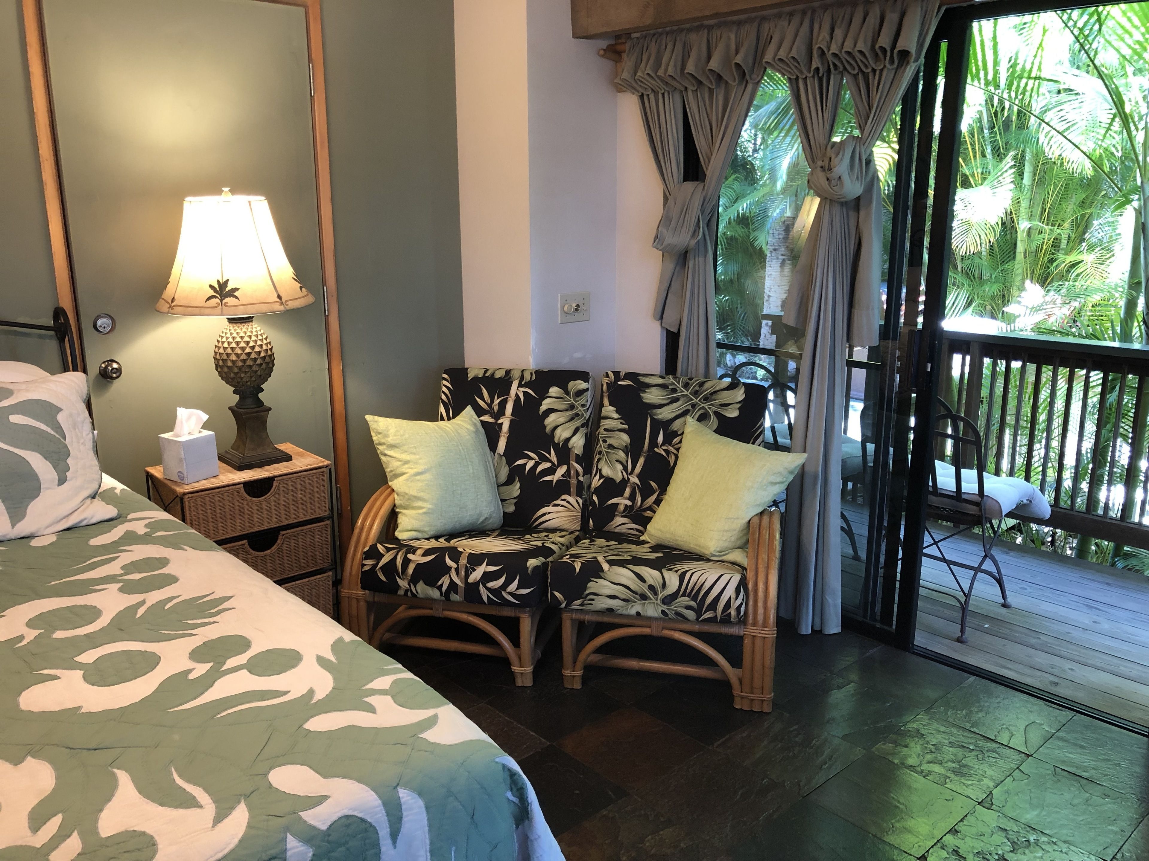 A room with a bed, a chair with cushions, a lamp, and a view of greenery through a window.