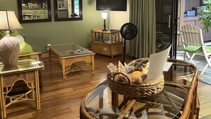 A living room with a glass-top table, wicker chairs, a ceiling fan, a flat-screen TV, and a sliding glass door leading to an outdoor area. - Maui What A Wonderful World B and B Hawaii  (Kihei)