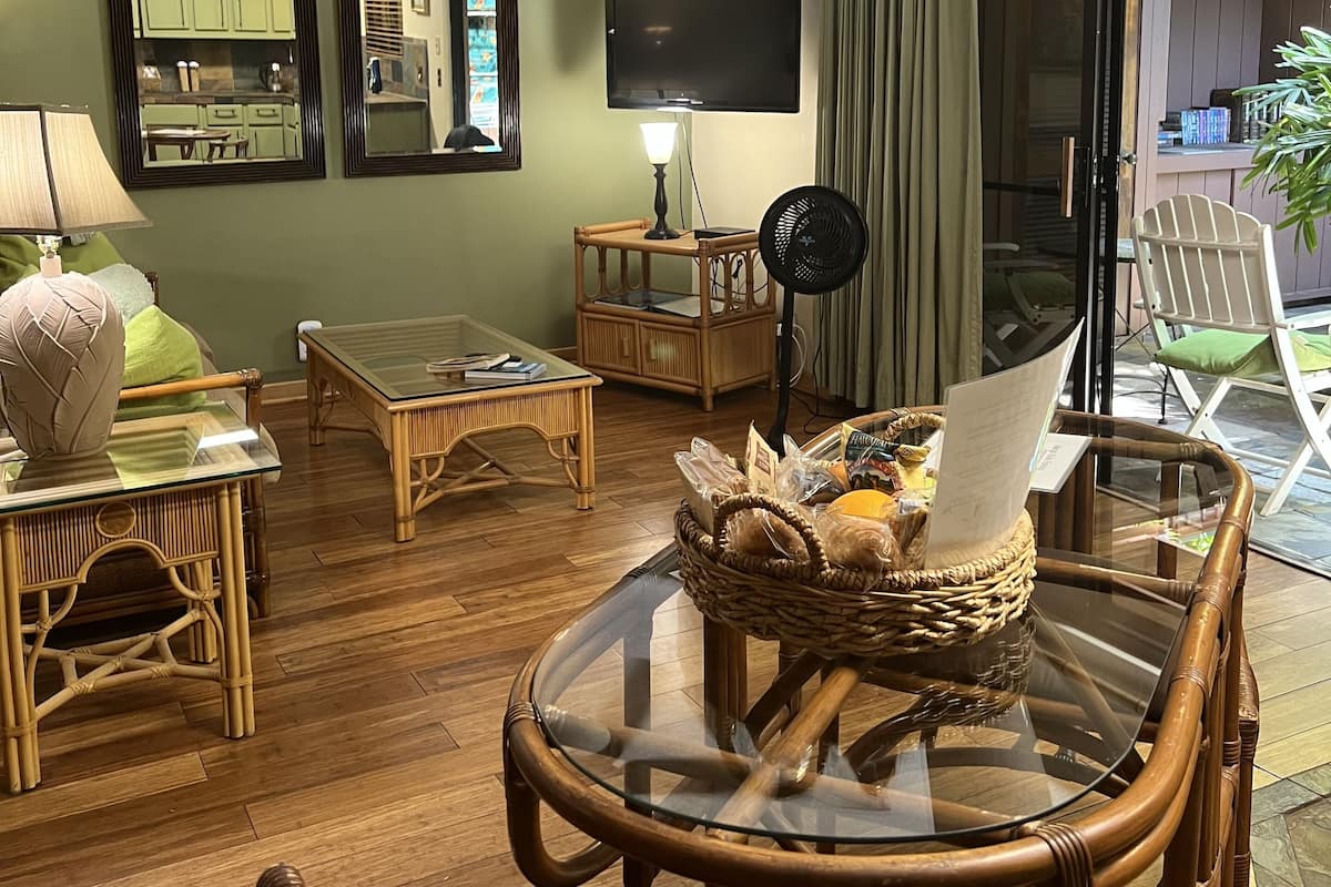 Un salon doté d’une table à plateau en verre, de fauteuils en osier, d’un ventilateur de plafond, d’un téléviseur à écran plat et d’une porte-fenêtre coulissante donnant sur un espace extérieur.