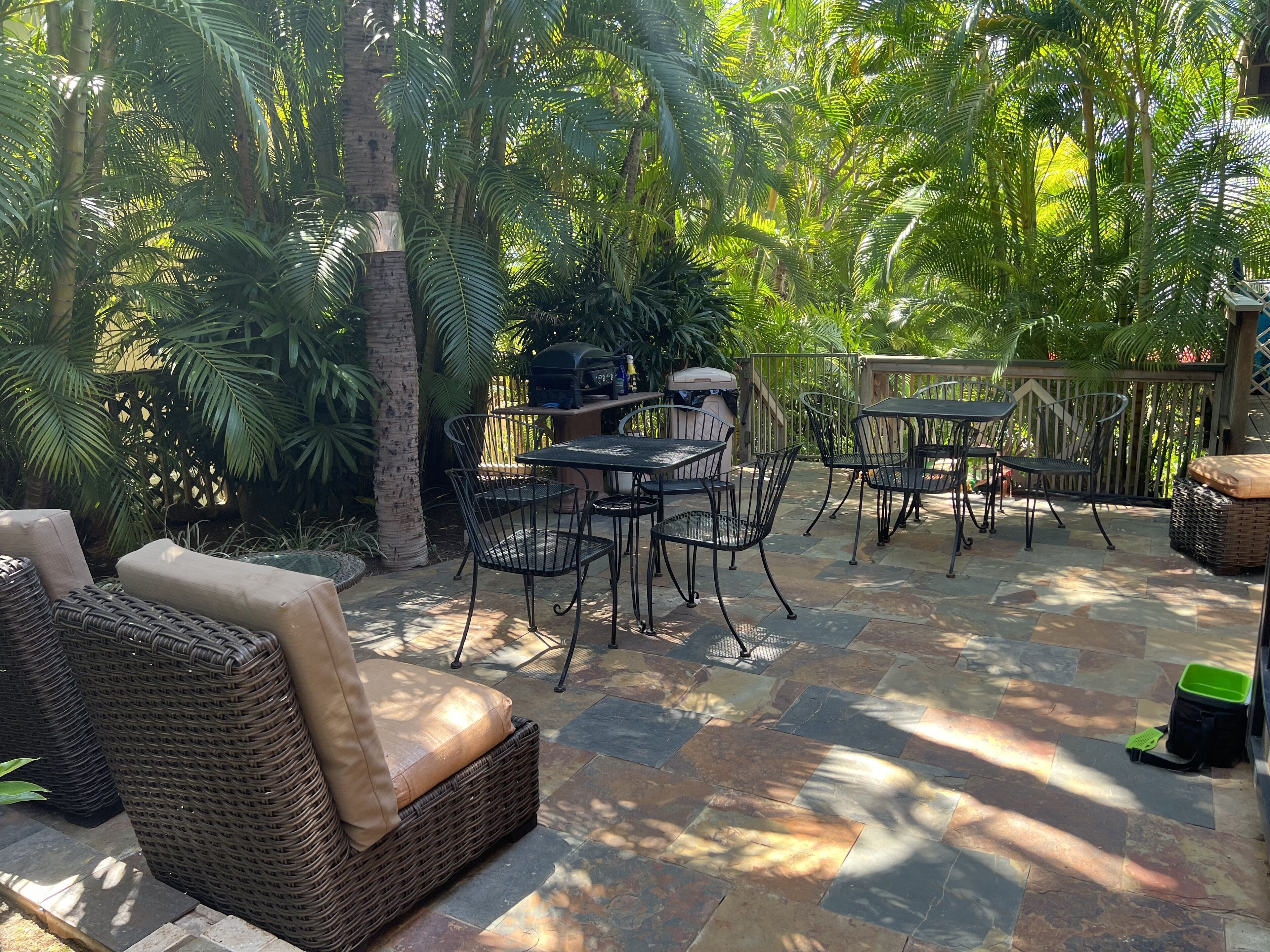 An outdoor patio with wicker furniture, stone flooring, and a grill area surrounded by lush greenery.