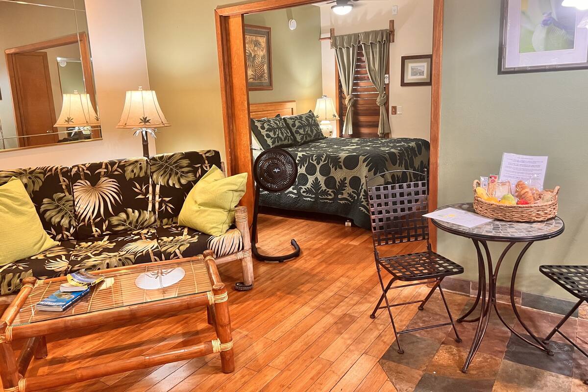 Une pièce avec un plancher en bois, un canapé orné d’un motif de feuilles, un lit orné d’un motif de feuilles, un ventilateur sur pied, une petite table avec un panier de fruits et un miroir accroché au mur.