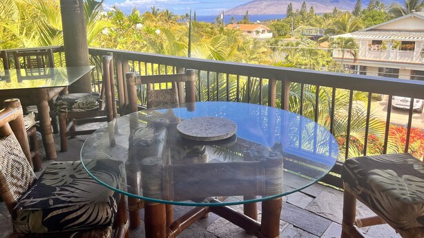 A covered outdoor area with a glass table and chairs, surrounded by lush greenery and a view of a mountain and buildings. - Maui What A Wonderful World B and B Hawaii  (Kihei)