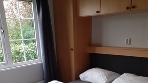 Una cama bien hecha con sábanas y almohadas blancas, una ventana con vista a la vegetación, y cabecera y mesita de noche de madera.