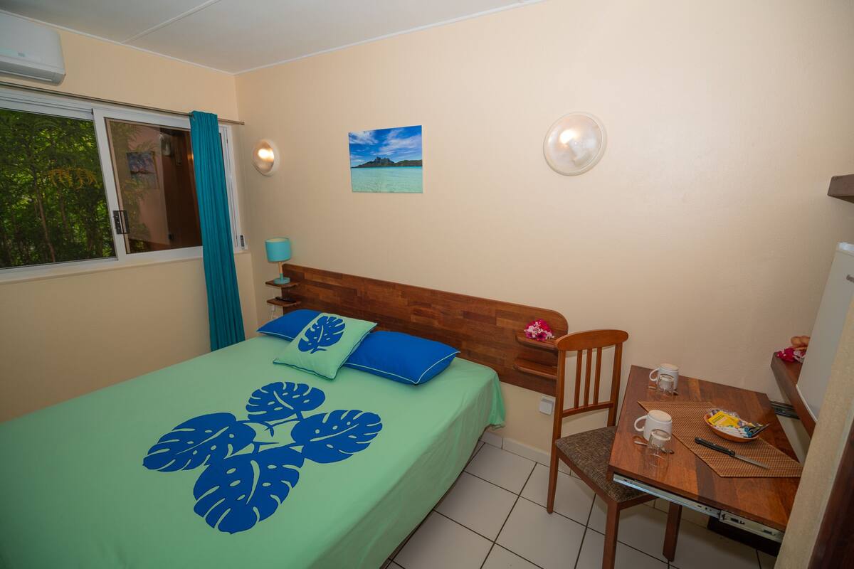 A bedroom with a bed, a wooden headboard, a bedside table with a lamp, a chair, and a small table with a bowl and a cup.
