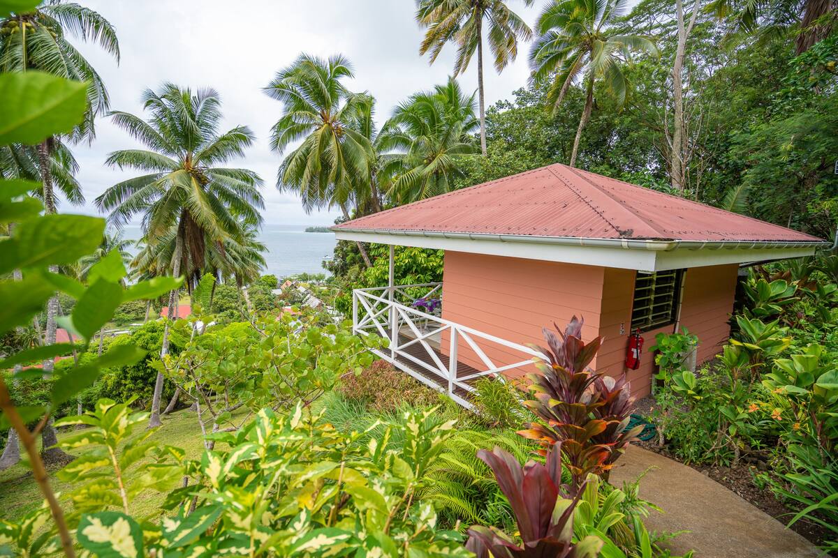 A small building with a red roof surrounded by lush greenery and palm trees, overlooking a body of water.