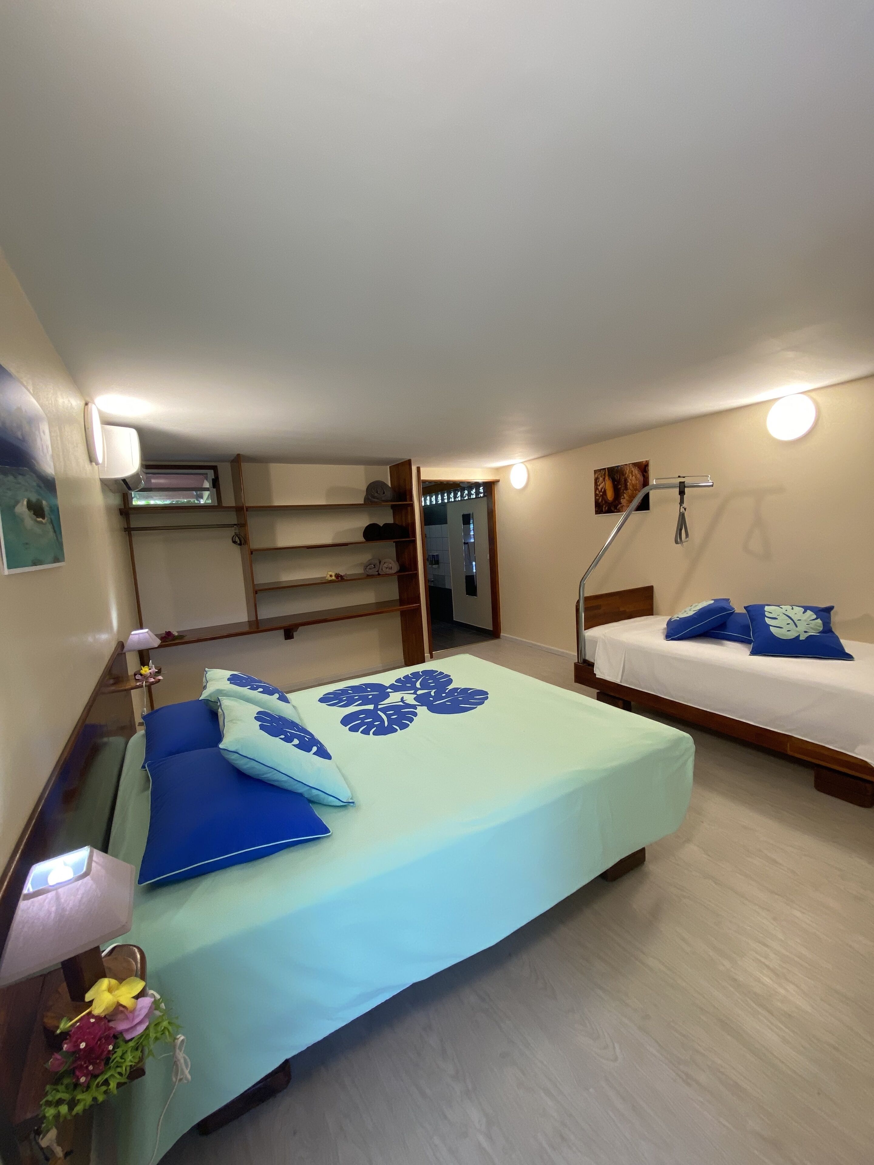 A bedroom with a bed, a bedside table, a lamp, a wall-mounted shelf, and a door leading to another room.