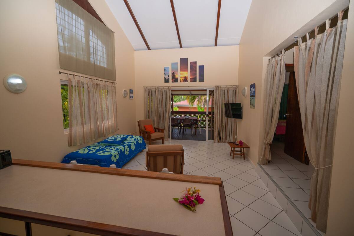 A hotel room with a bed, a chair, a small table, and a TV. There is a balcony with a view of the outside.