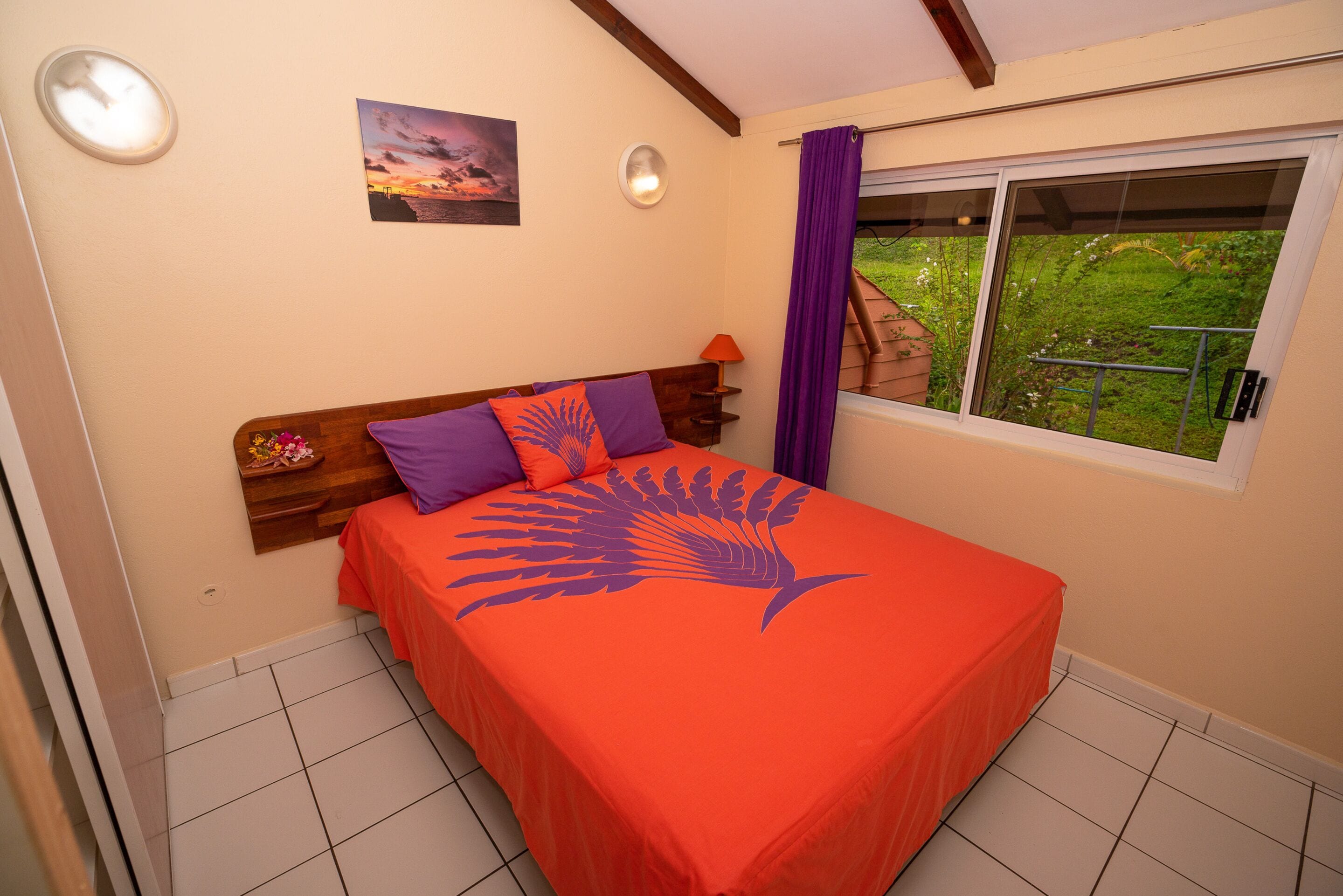 Un dormitorio con cama, una mesita de noche, una ventana con vistas a la vegetación y un cuadro en la pared.