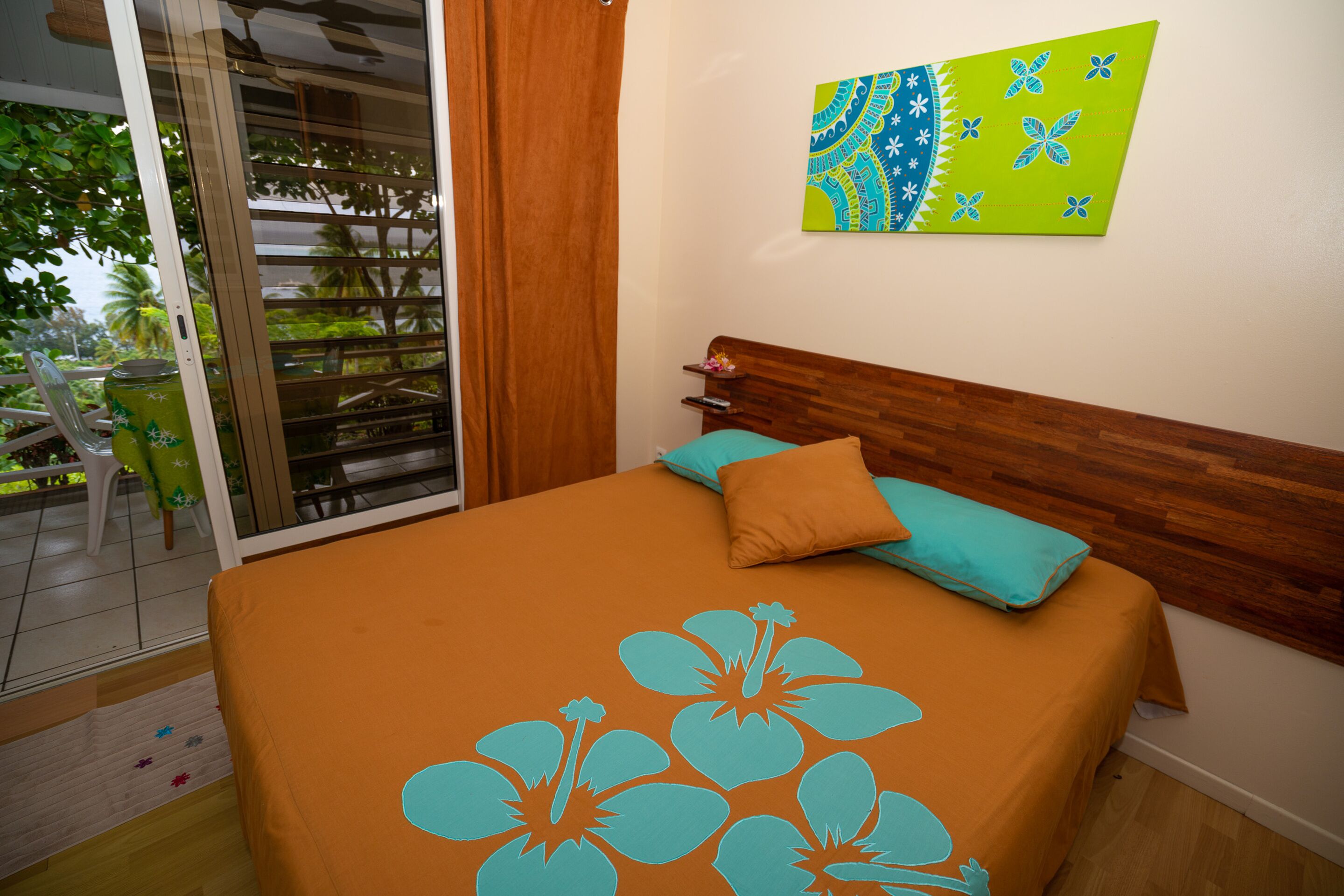 Une chambre à coucher avec une tête de lit en bois, un lit recouvert d’une couverture orange à motifs floraux bleus, un coussin vert et une peinture abstraite colorée accrochée au mur.