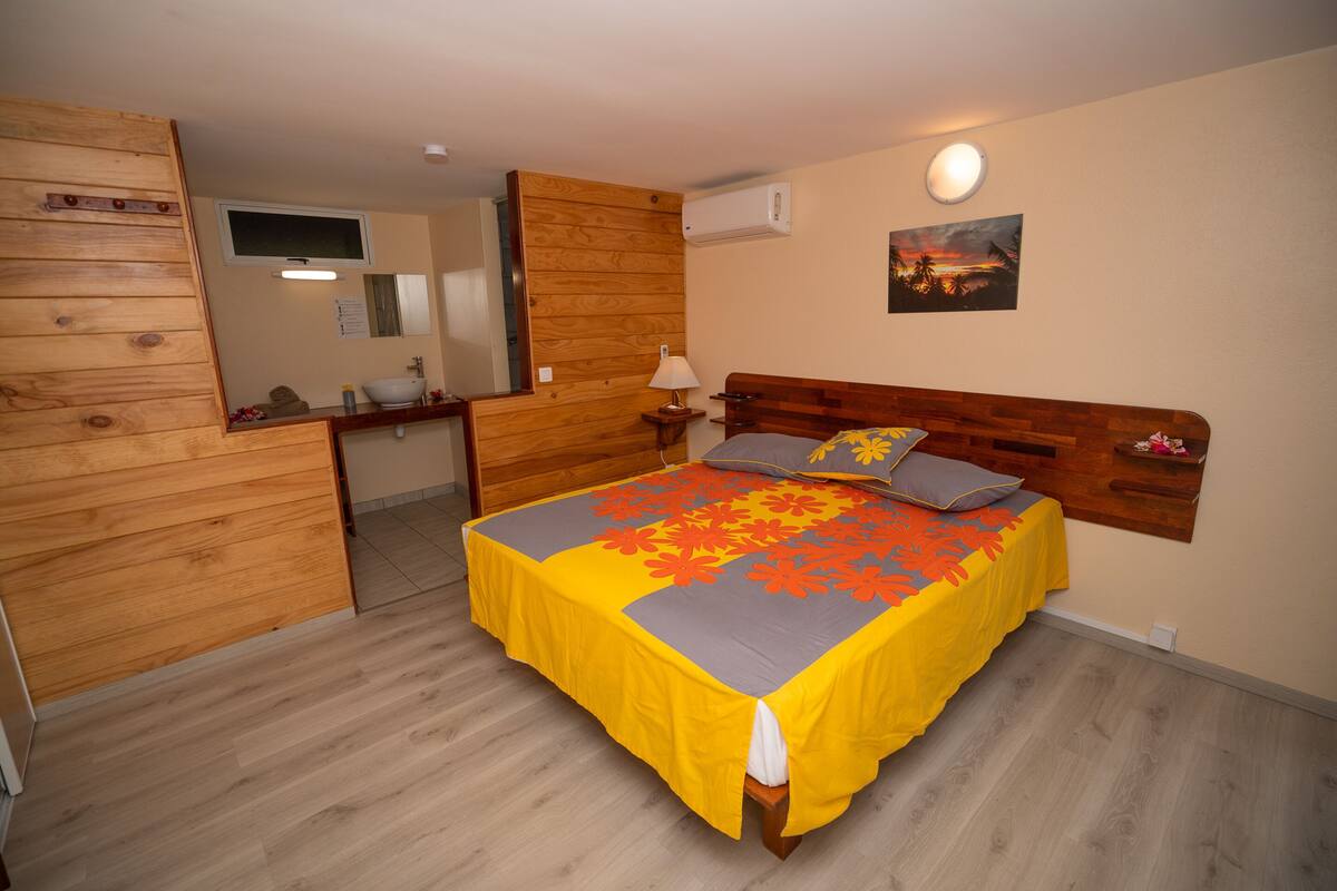 A bedroom with a wooden headboard, a bed with a yellow and gray floral bedspread, a nightstand with a lamp, and a wall-mounted TV.