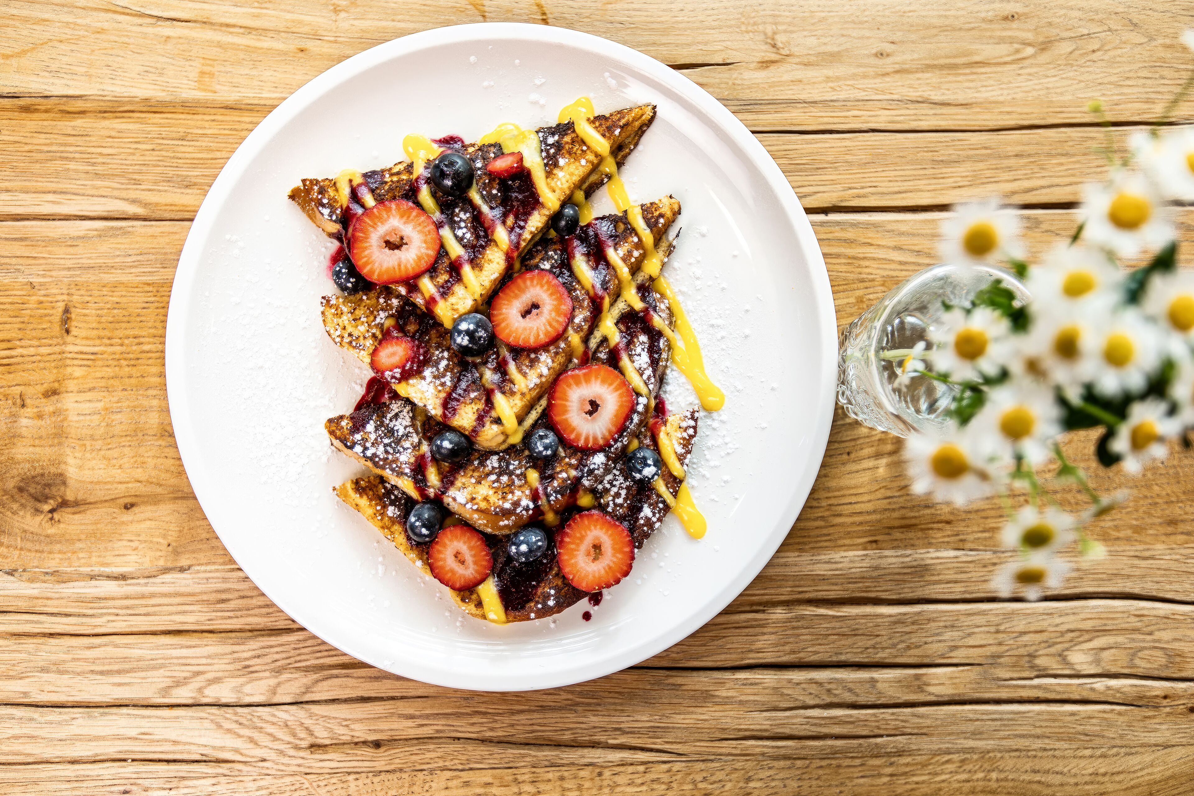 A plate of French toast with strawberries and blueberries, dusted with powdered sugar, on a wooden table.