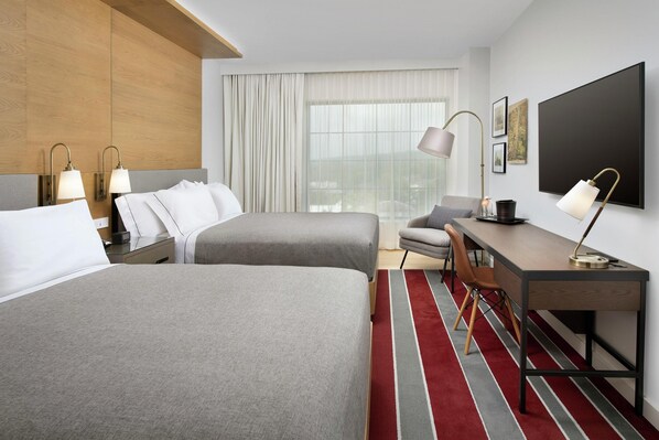 Habitación de hotel con dos camas, un escritorio, una silla, un televisor y un ventanal grande con cortinas.