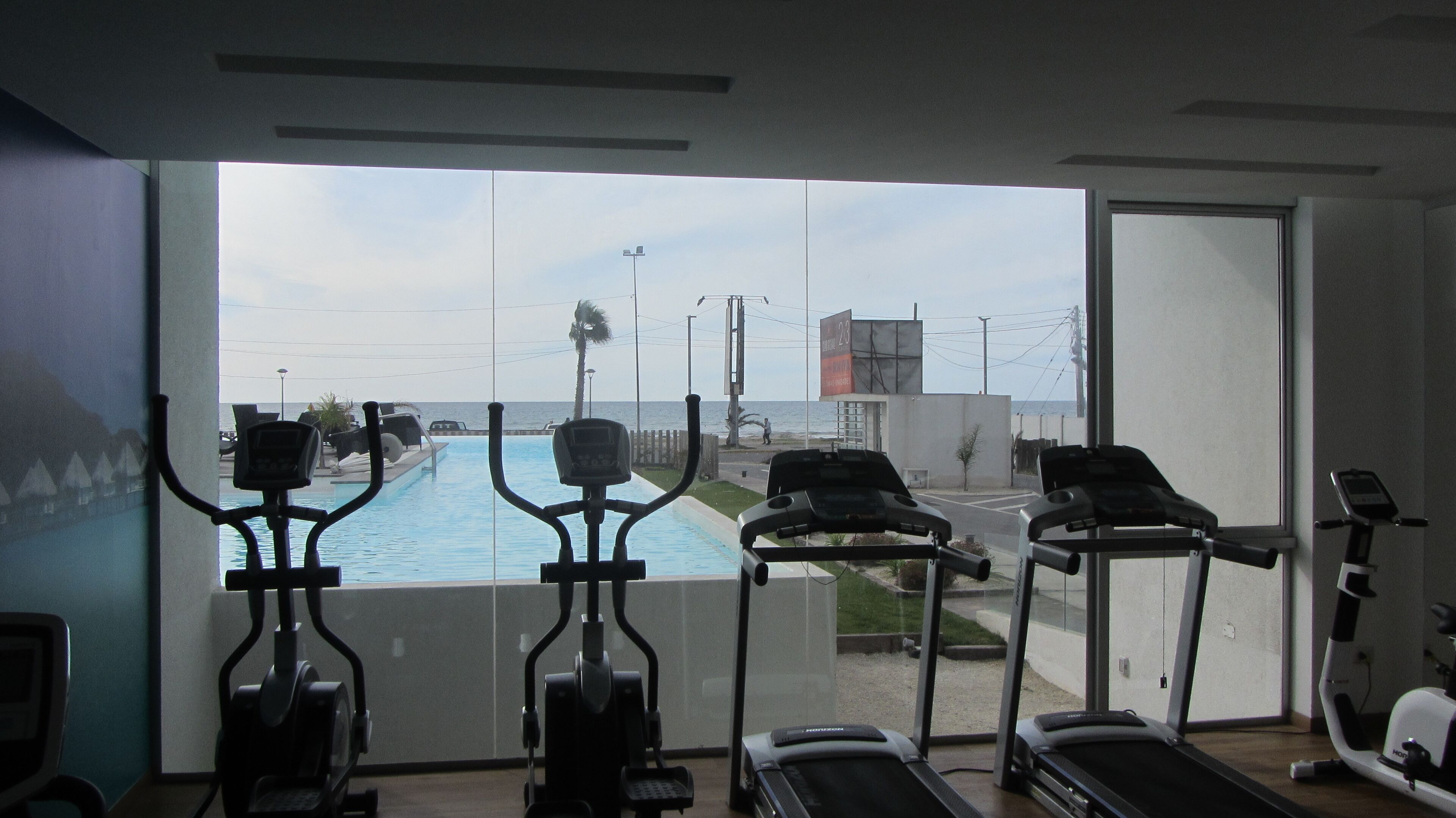 Un gimnasio con cintas de correr y una máquina elíptica, con vistas a una piscina y a una calle con vista al océano.