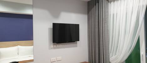 Habitación de hotel con televisor de pantalla plana en la pared, mesita de noche de madera con tetera y tazas, espejo y ventana con cortinas grises.