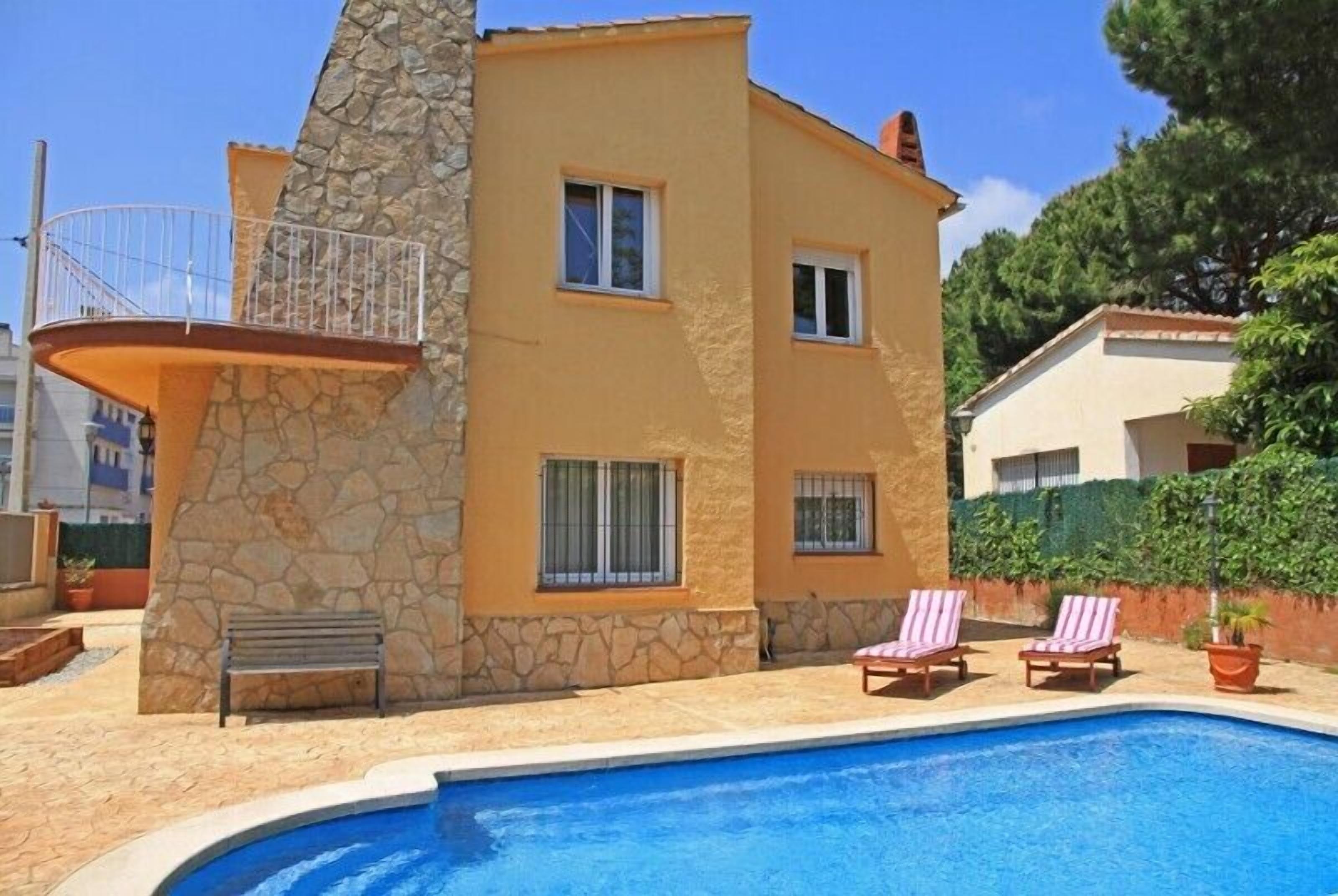 A two-story building with a stone facade, a swimming pool, and two lounge chairs.