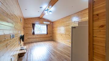 Habitació àmplia amb parets i terra de fusta. Hi ha una finestra amb cortines, una làmpada al sostre i un frigorífic.