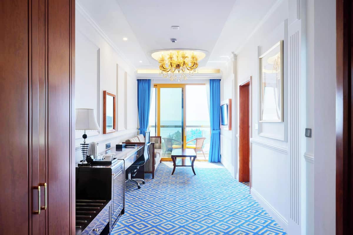 A hotel corridor with a patterned floor, a chandelier, a desk with a lamp, a sofa, and a view of the outdoors.