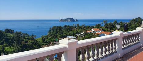 A balcony with a stone balustrade overlooking a coastal landscape with houses and a cliff.