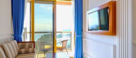A room with a sofa, a table, a TV mounted on the wall, and a balcony with a view.