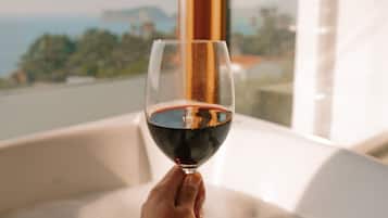Una mano sostiene una copa de vino tinto, colocada sobre una bañera con vistas a un paisaje a través de una ventana.