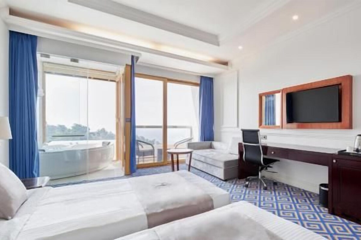 A hotel room with a bed, a desk, a chair, a TV, and a balcony with curtains.