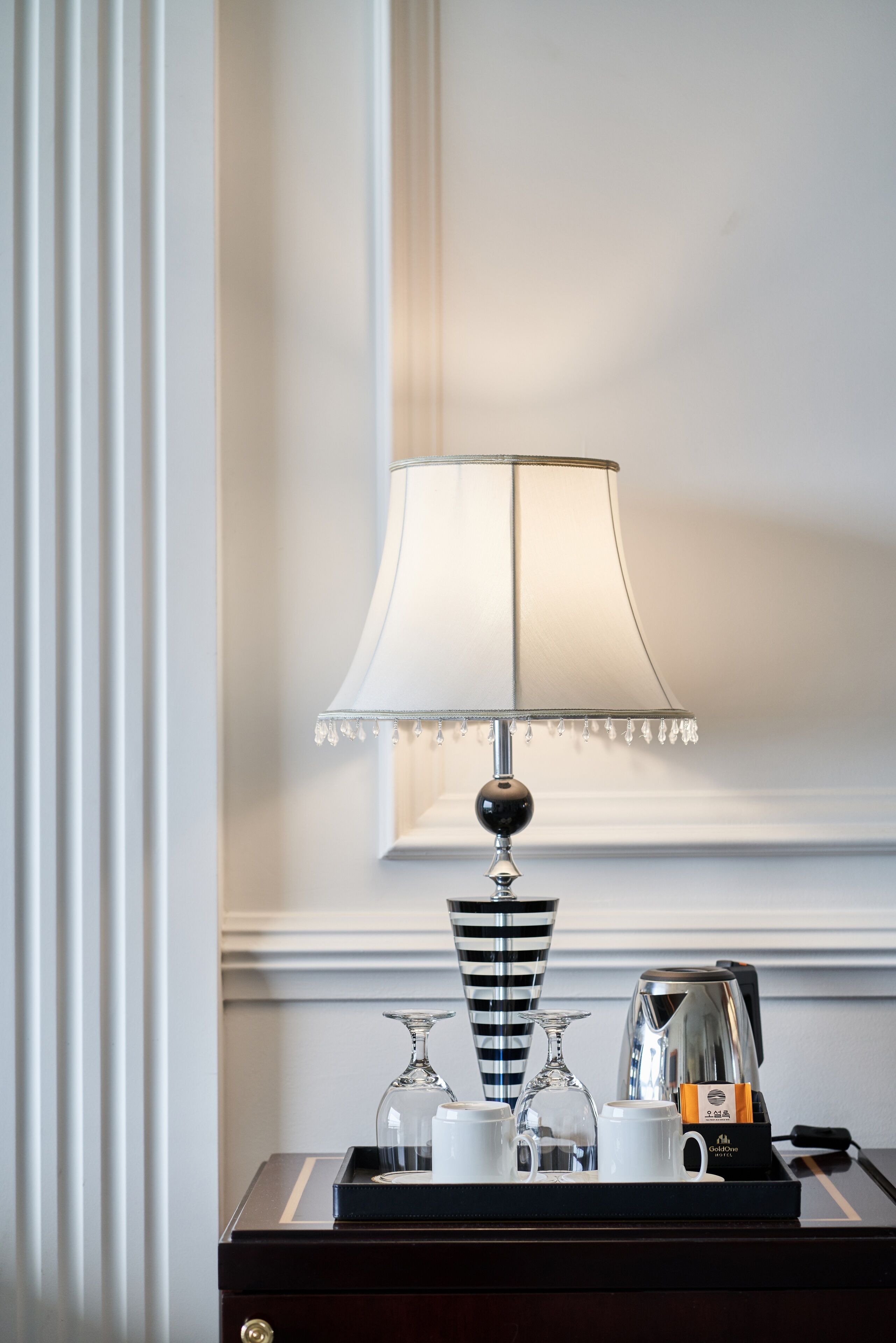 A hotel room with a bedside table featuring a lamp, a tray with cups, a glass, and a coffee maker.