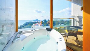A modern bathroom with a large bathtub, a glass shower enclosure, and a view of a coastal landscape. - GoldOne Hotel & Suites (Seogwipo)