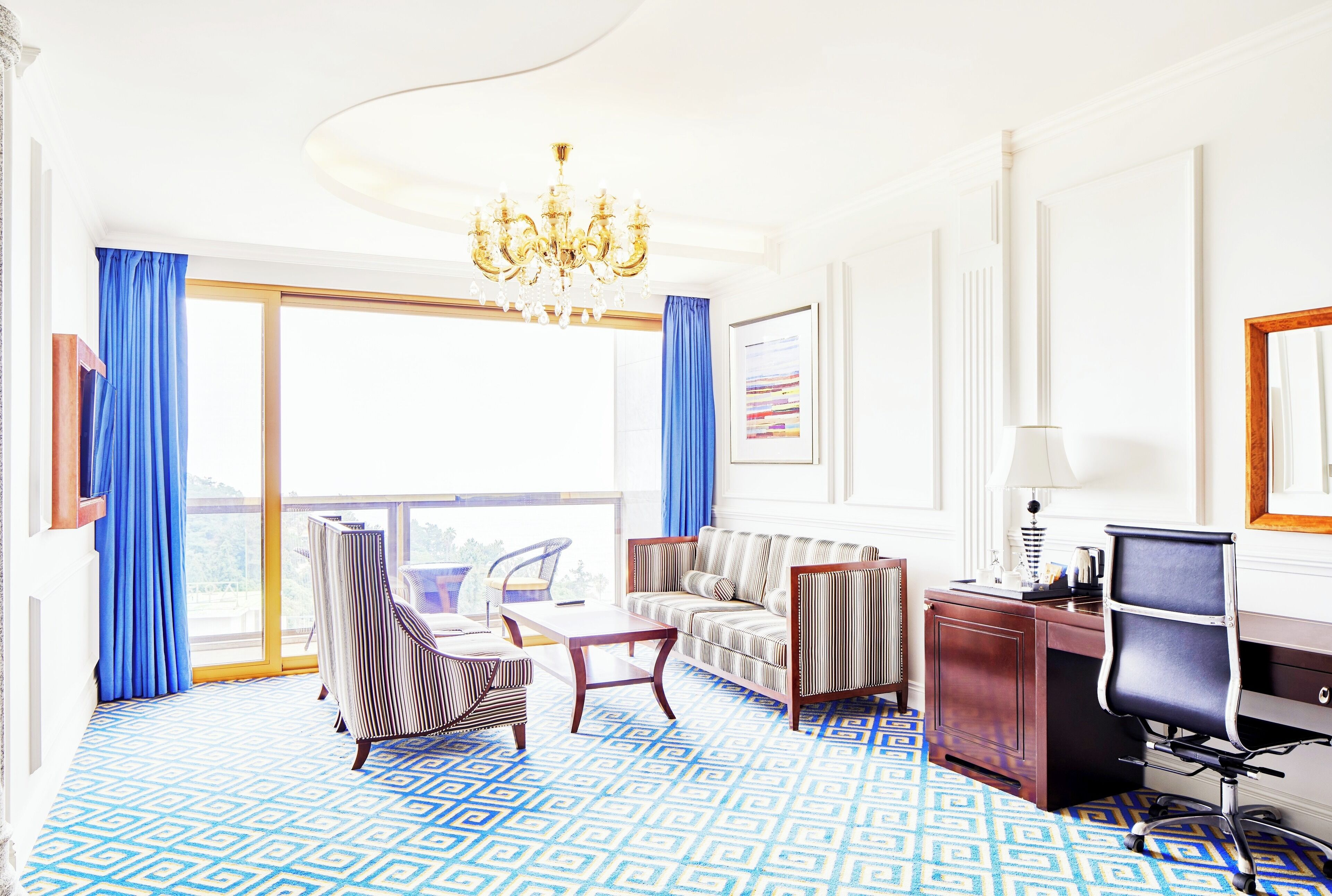 A hotel room with a large window, a chandelier, a sofa, a desk, and a chair.