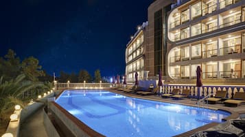 A nighttime pool area with lounge chairs and umbrellas, surrounded by a building with multiple balconies and large windows.
