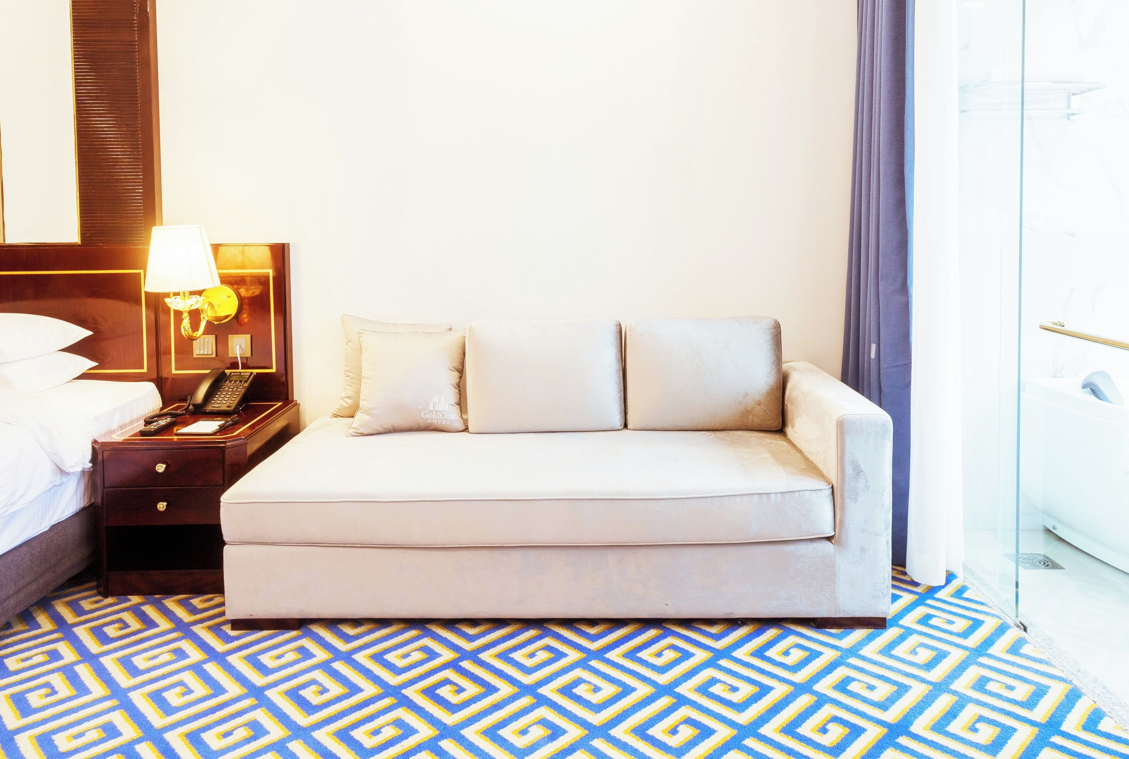 A hotel room with a bed, a nightstand with a lamp, a sofa, and a patterned carpet.