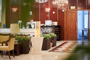 A lobby with a checkered floor, a reception desk, a lamp, chairs, and potted plants.
