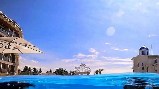 A swimming pool with clear blue water, a tiled floor, and a building with a dome and balcony in the background. - GoldOne Hotel & Suites (Seogwipo)