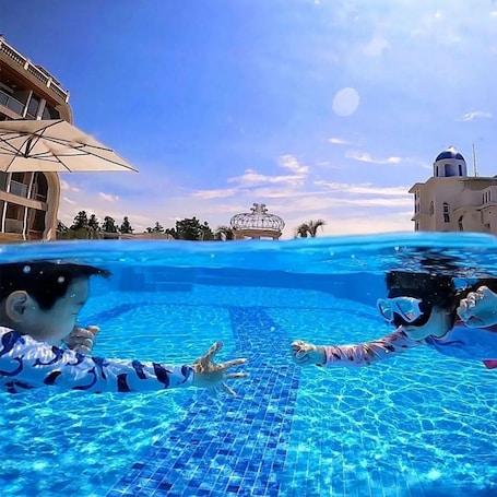Una piscina con agua azul cristalina, piso de baldosas y un edificio con cúpula y balcón al fondo.
