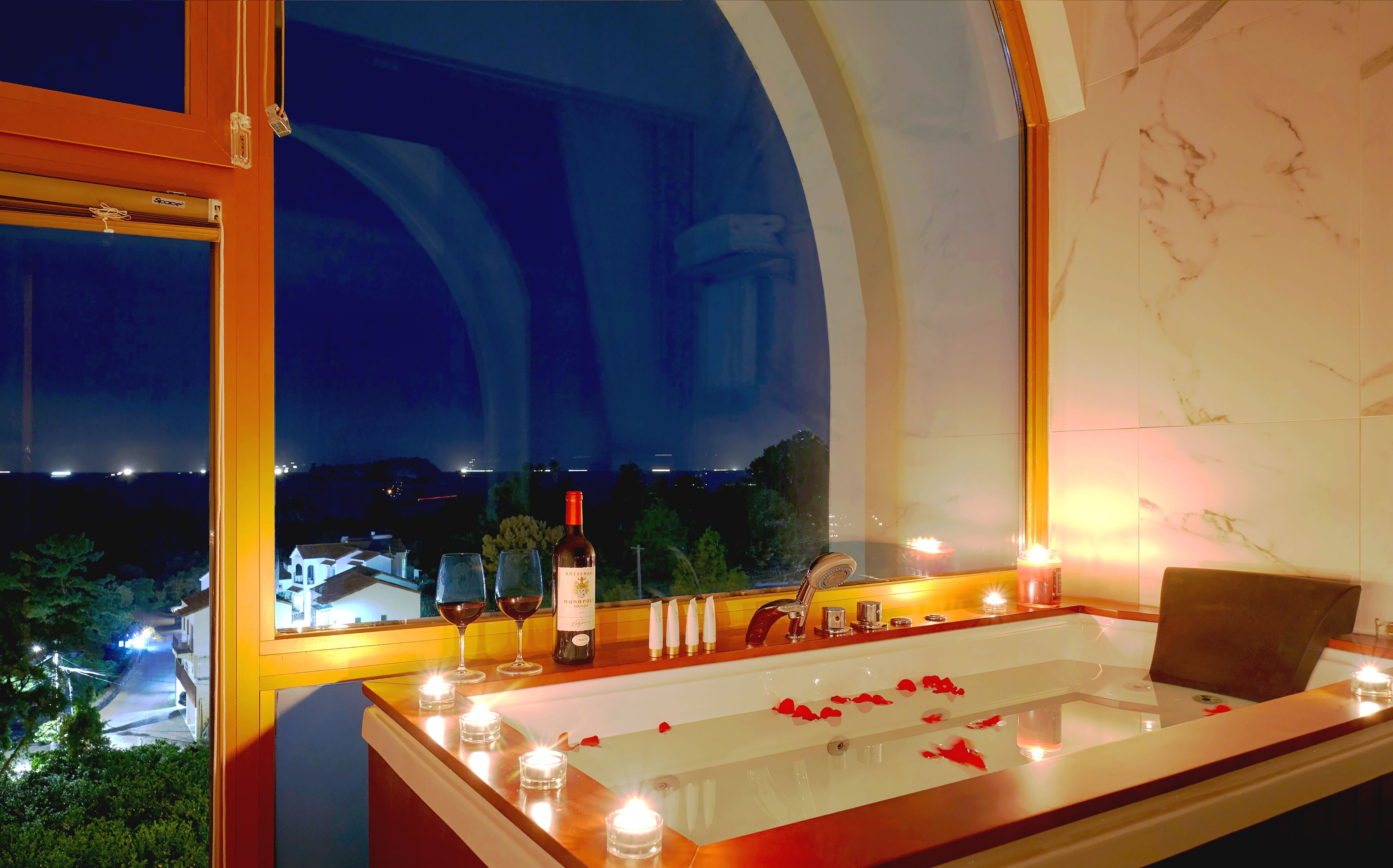 A bathtub with rose petals, candles, and a bottle of wine, with a view of a cityscape at night.