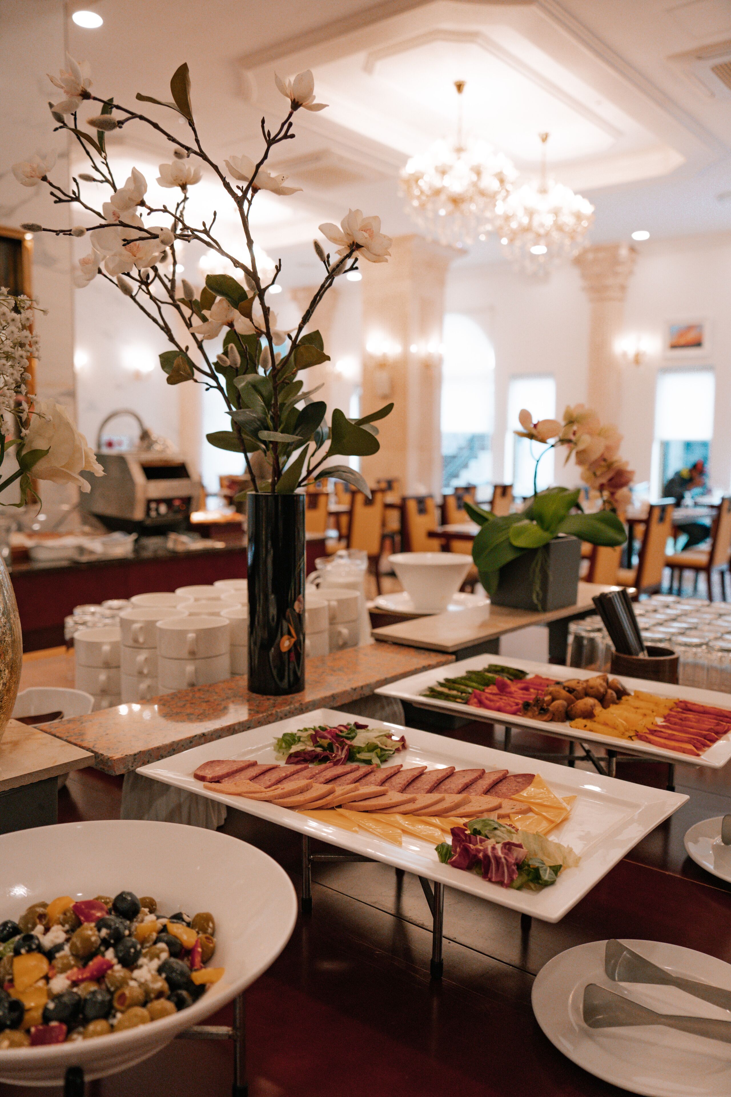 Una mesa de buffet con diversos alimentos, entre ellos ensalada, queso y carnes, en un comedor bien iluminado con candelabros y vistas a la ciudad.