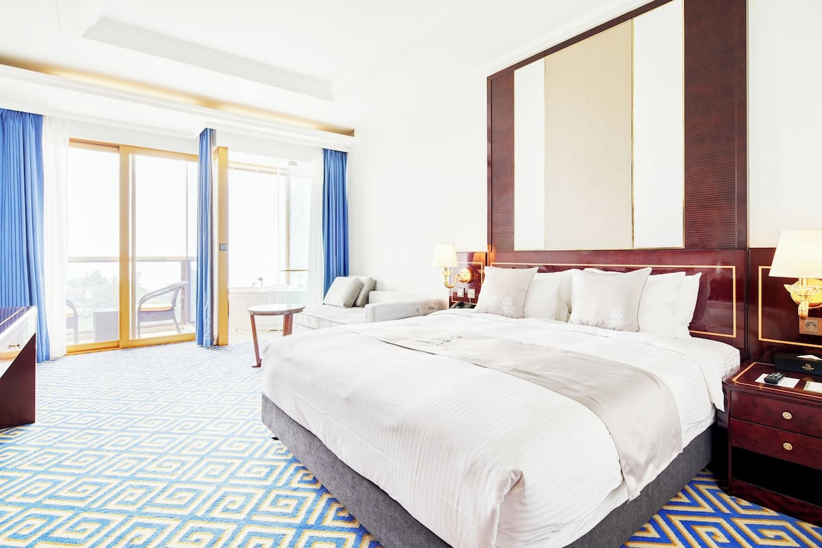 A hotel room with a large bed, a desk, a chair, a nightstand, and a large window with curtains.