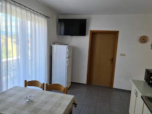 Une pièce comprenant une table à manger, des chaises, un téléviseur fixé au mur, une porte en bois et un coin cuisine.