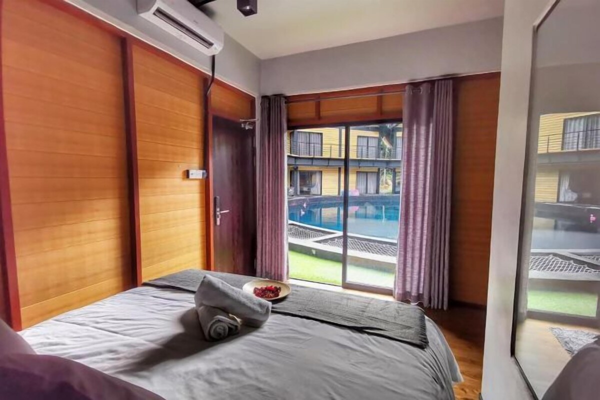 Habitación de hotel con cama, una mesita con un plato de comida, una puerta corredera y vistas a una piscina y edificios.