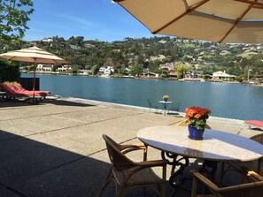 A patio with lounge chairs and a table with a vase of flowers, overlooking a lake with houses in the background. - New, completely rebuilt  two level Belvedere Lagoon waterfront monthly rental.  (Belvedere)
