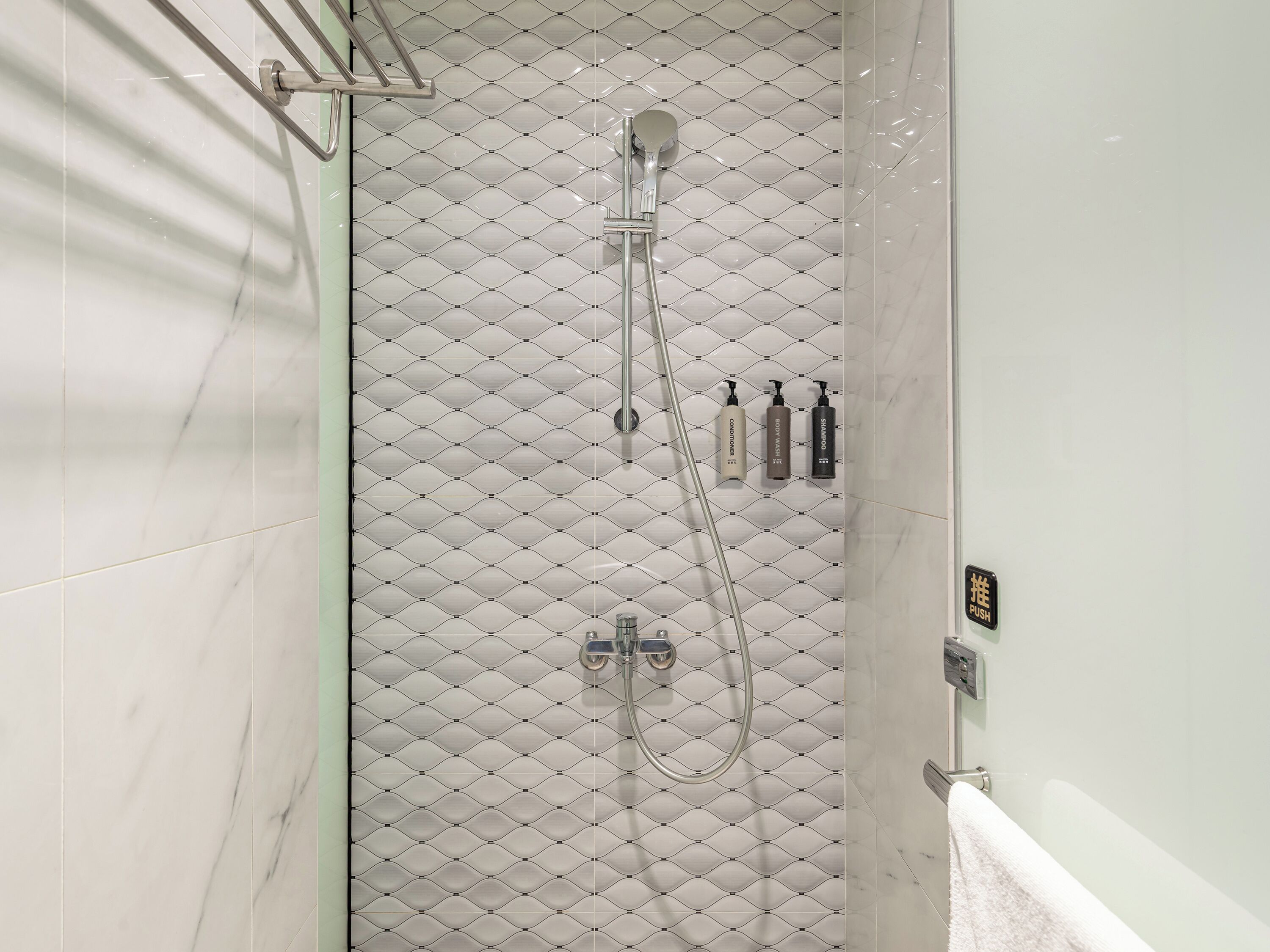 Une douche avec un mur recouvert de carreaux disposés en motif losange, un pommeau de douche à main et trois bouteilles de shampoing.
