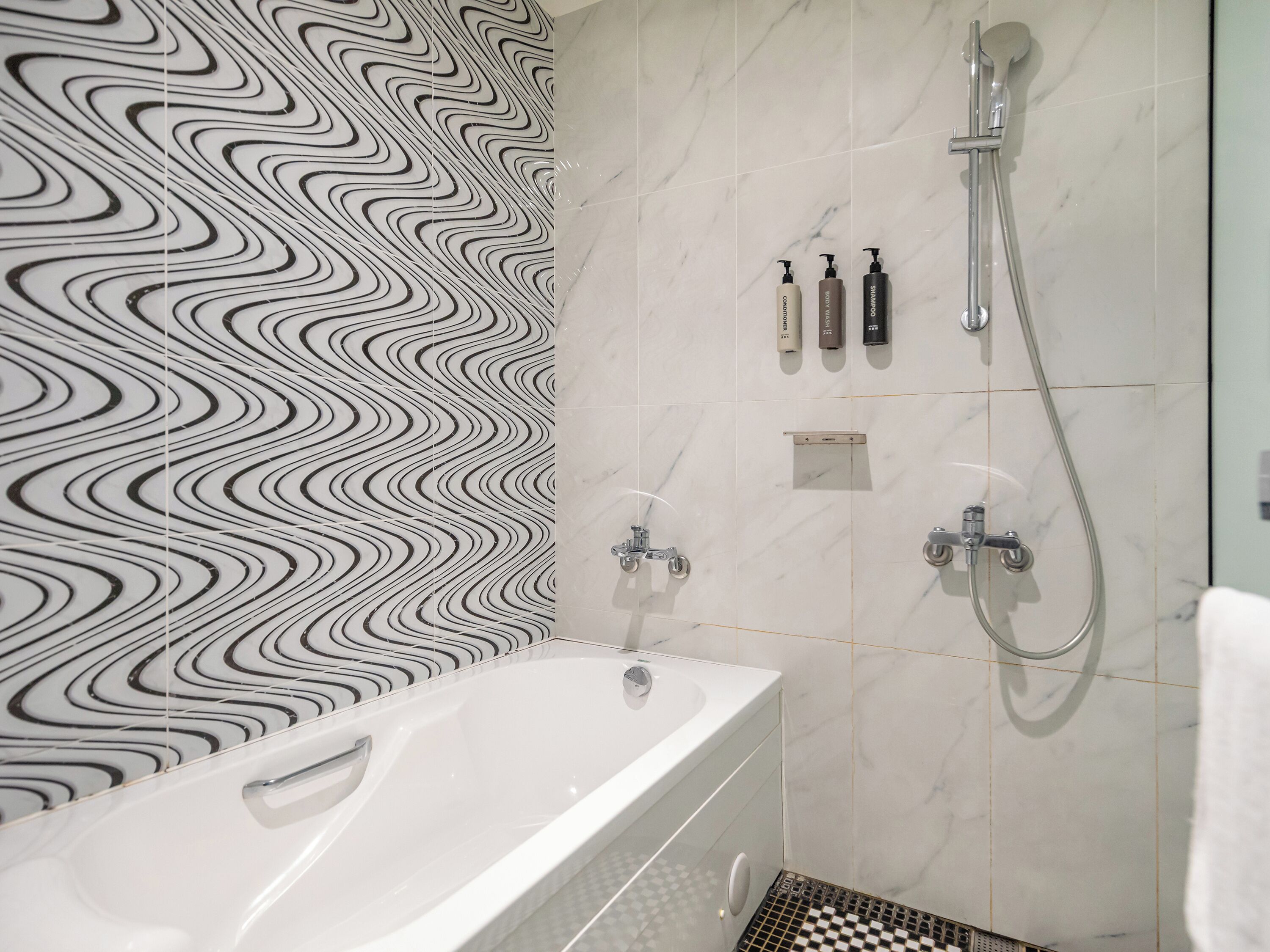 A bathroom with a white bathtub, a shower with a handheld showerhead, and a tiled wall with a black and white wavy pattern.