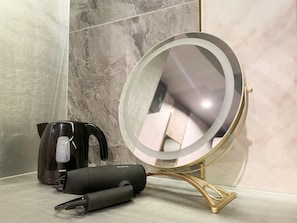 A bathroom with a marble wall, a black electric kettle, a compact mirror with a gold frame, and a black hair straightener.
