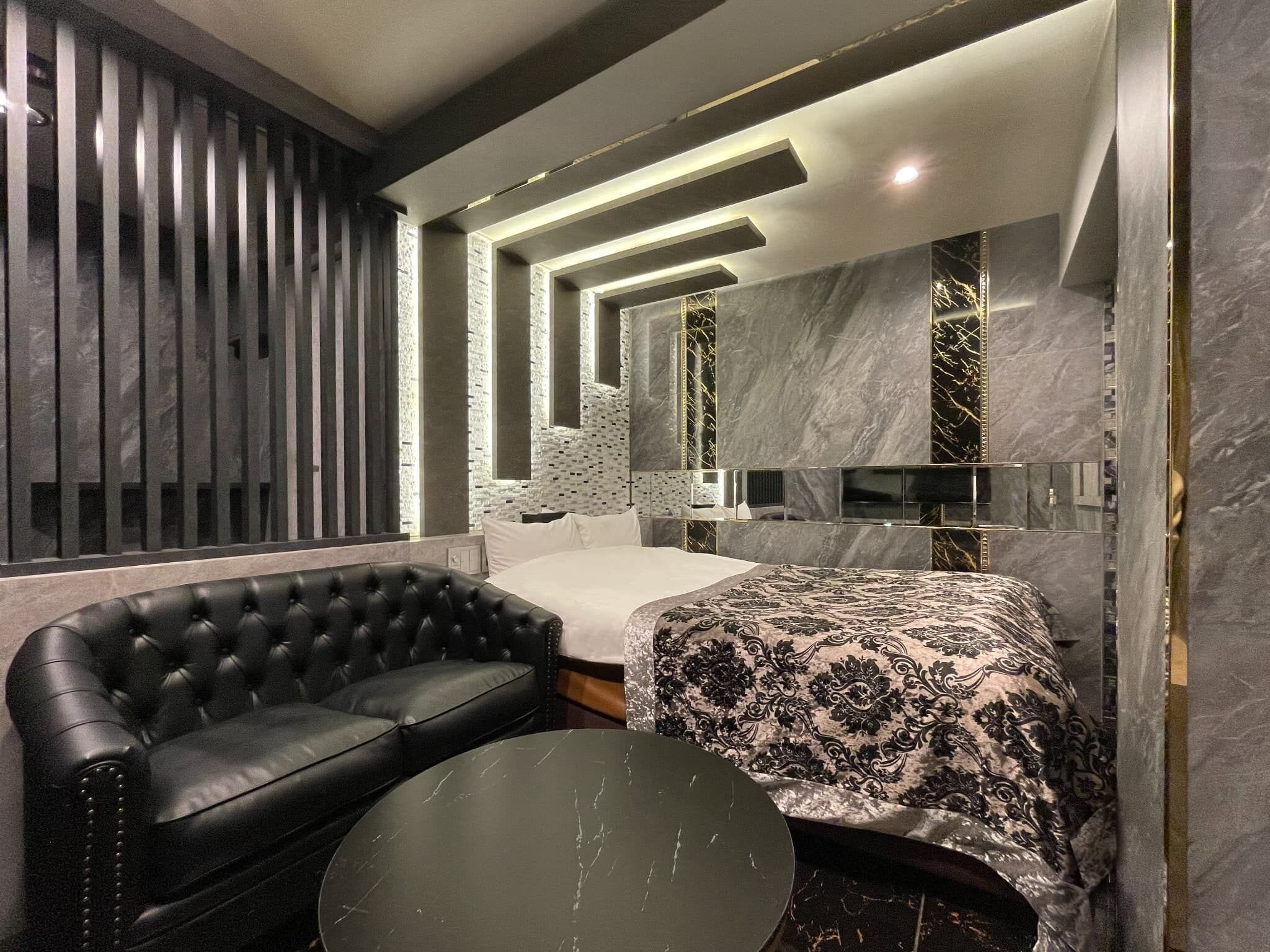 A modern bedroom with a black leather sofa, a round coffee table, a bed with a patterned bedspread, and a large mirror.