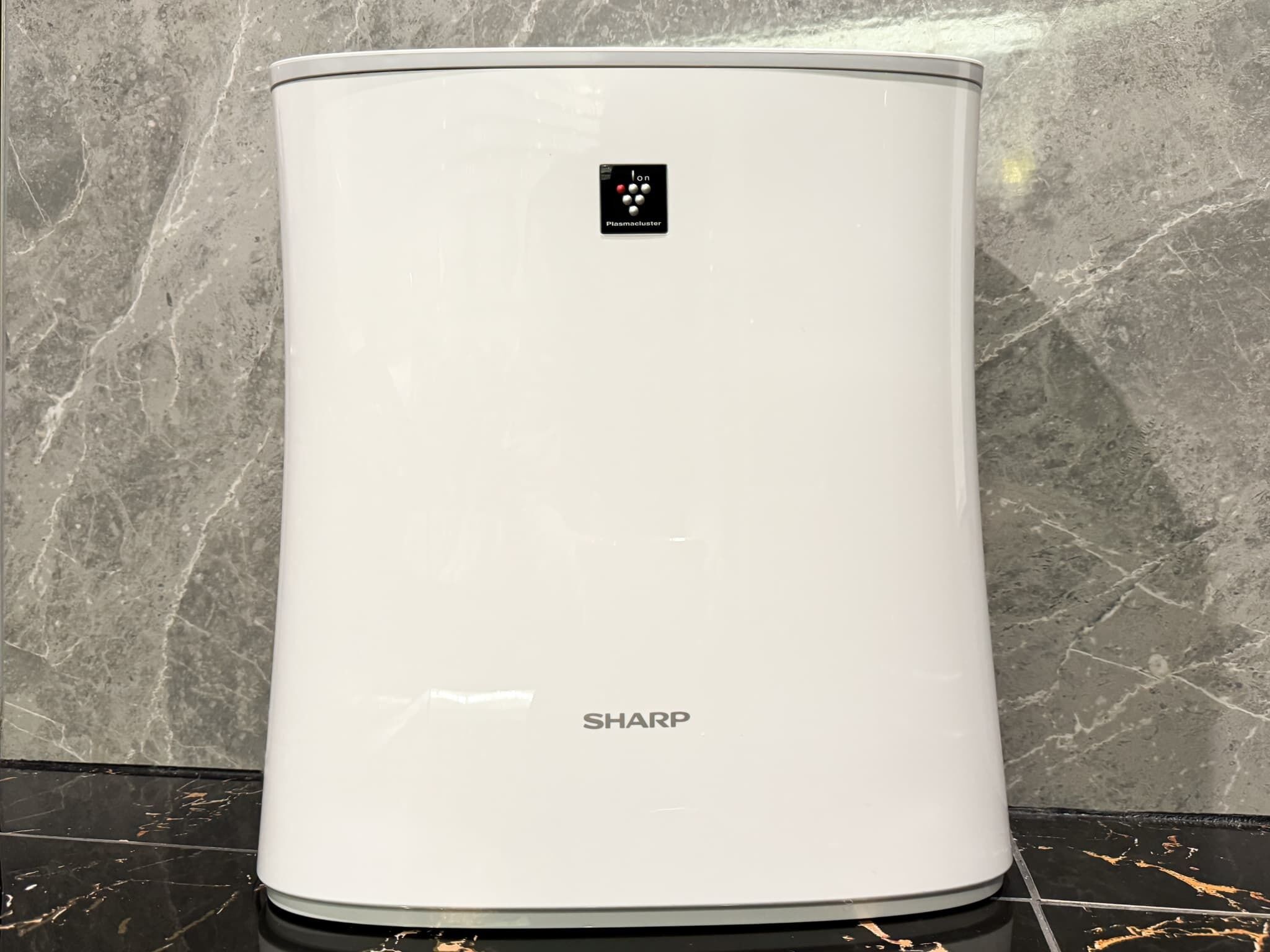 Calentador de agua SHARP blanco con panel de control, sobre un piso de baldosas negras frente a una pared de mármol.