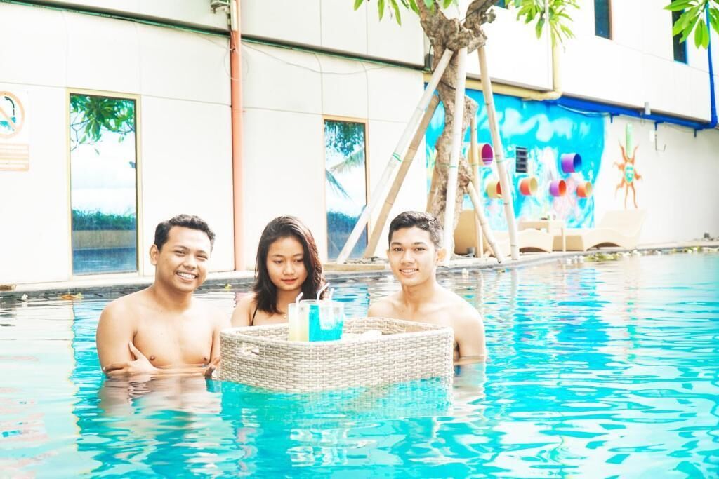 A swimming pool with three people, a wicker basket with drinks, and a building with a mural in the background.