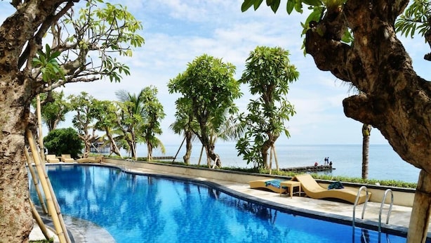 Una piscina azul cristalina rodeada de palmeras y sillas de descanso, con vista al mar.