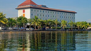 Un hotel grande con techo rojo y varios pisos, ubicado junto a una masa de agua con palmeras y coches estacionados.
