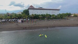 Un hotel frente a la playa con un edificio grande, palmeras y kayaks en el agua.