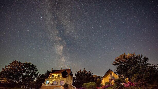 Una casa con la Vía Láctea visible en el cielo nocturno, rodeada de abundante vegetación y plantas con flores.